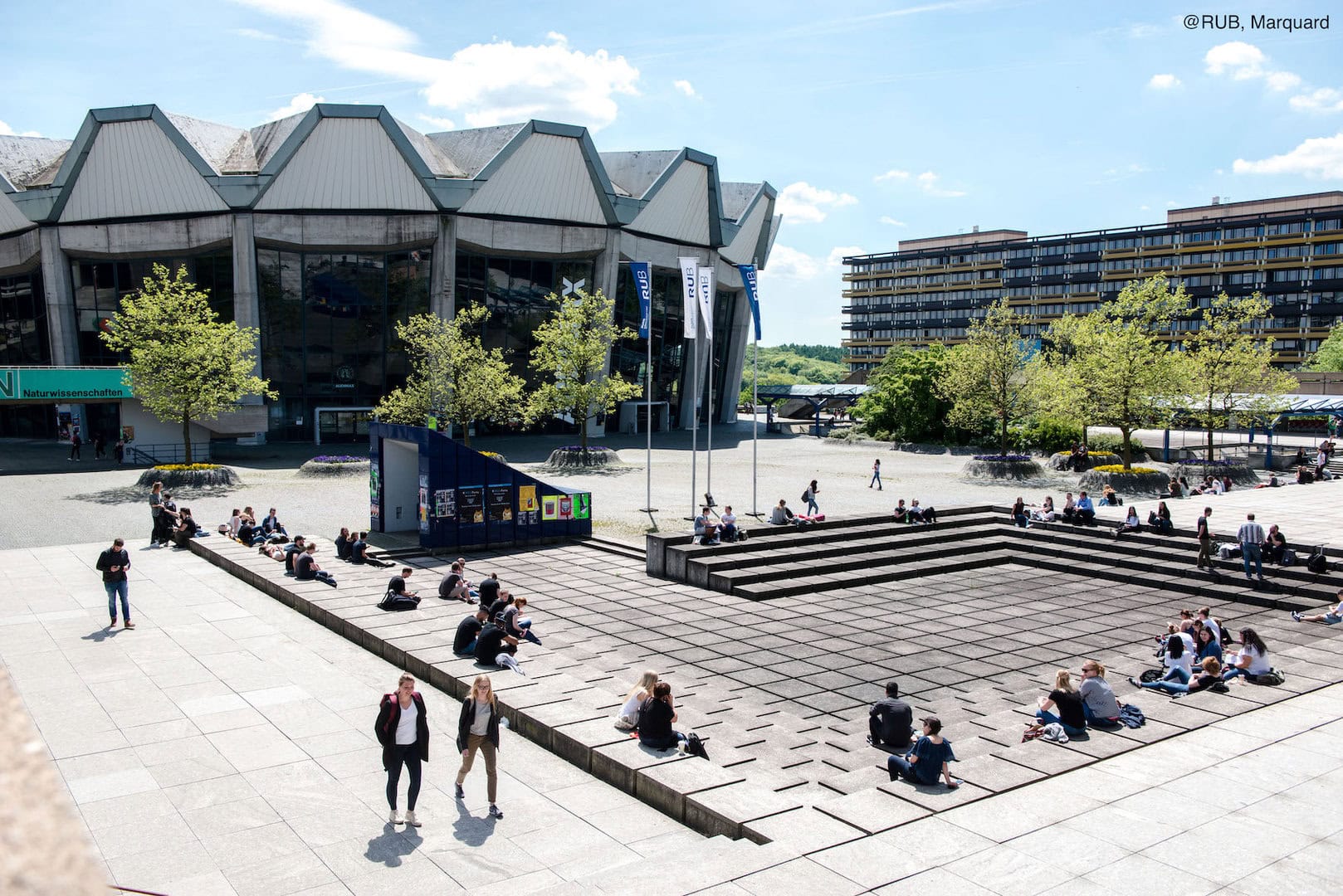 Menschen sitzen und spazieren im Freien vor einem großen, modernen Gebäude mit Glasfronten beim 2. Internationalen Kongress der IGSP.